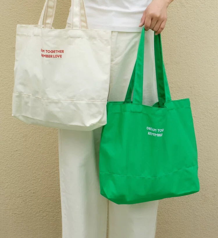 Person holding a white tote bag and a green tote bag with text on a beige background
