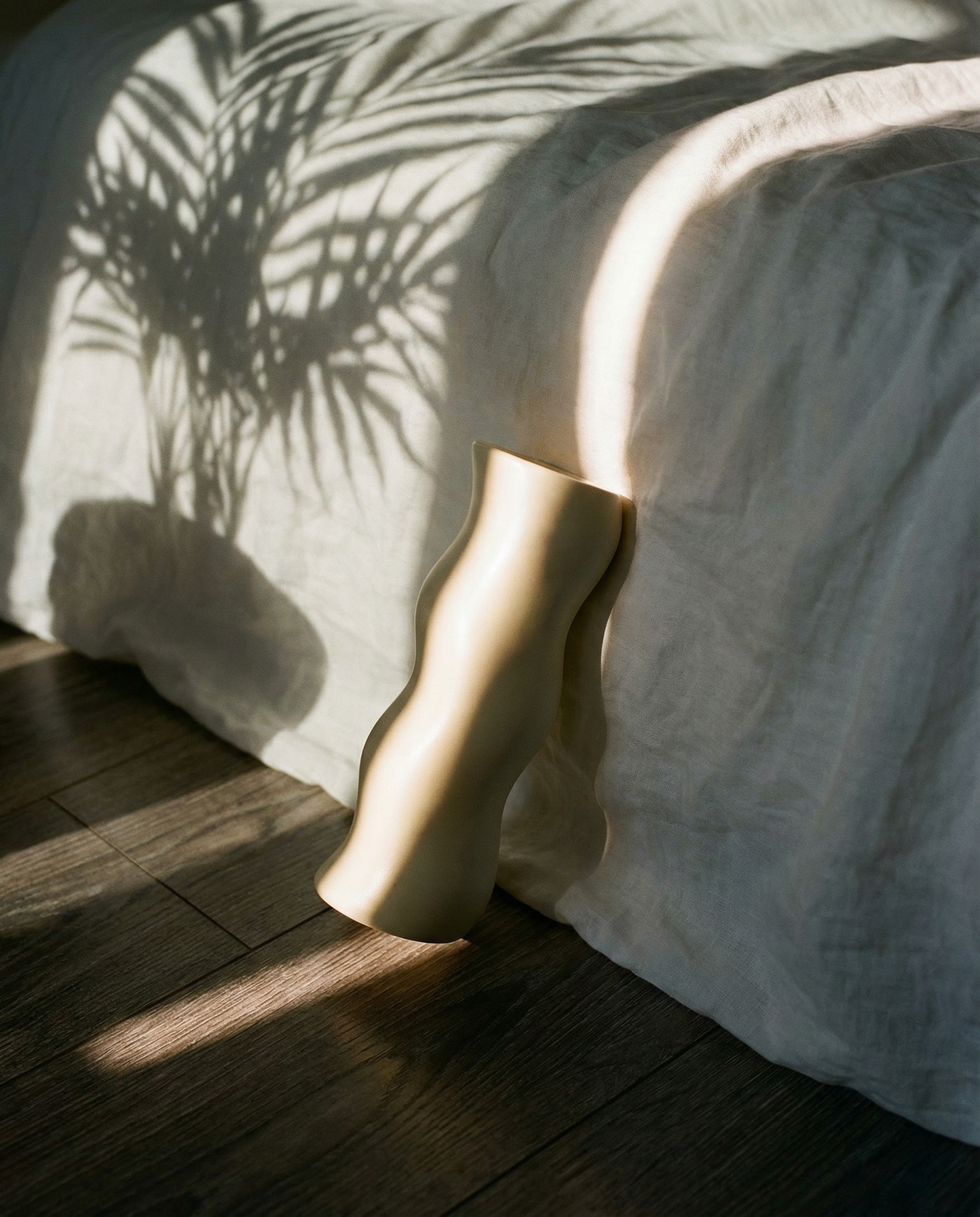 Vase on a wooden floor casting shadows on a white bedspread